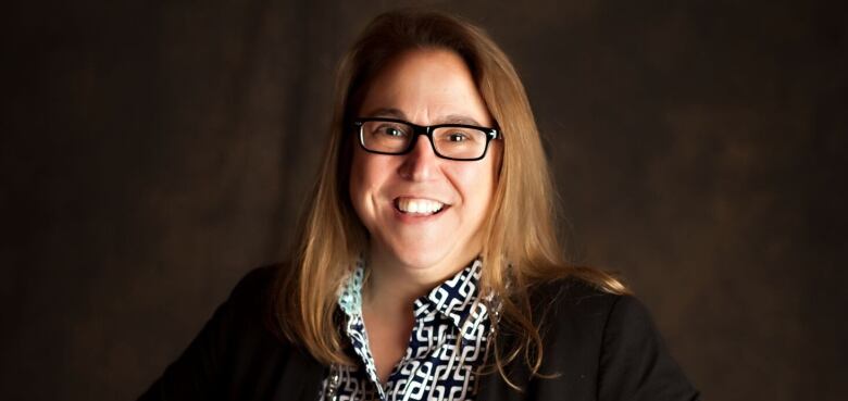 A woman with long blonde hair, dark rimmed glasses, wearing a black and white patterned shirt under a dark blazer.