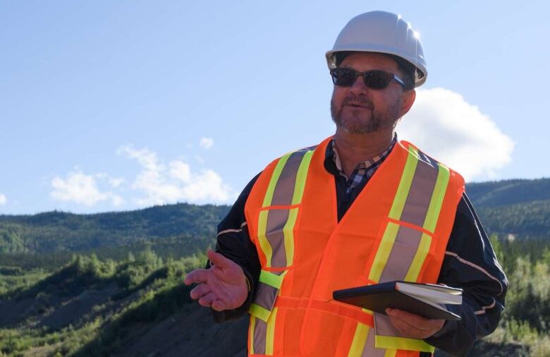 A man in a safety vest and hardhat stands outside in a remote, hilly area.