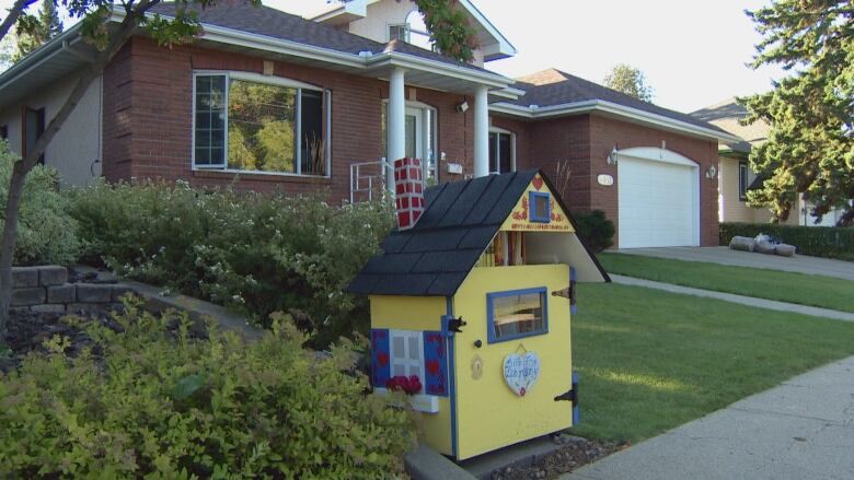 A yellow box for books next to a sidewalk. The box is decorated to look like a house.