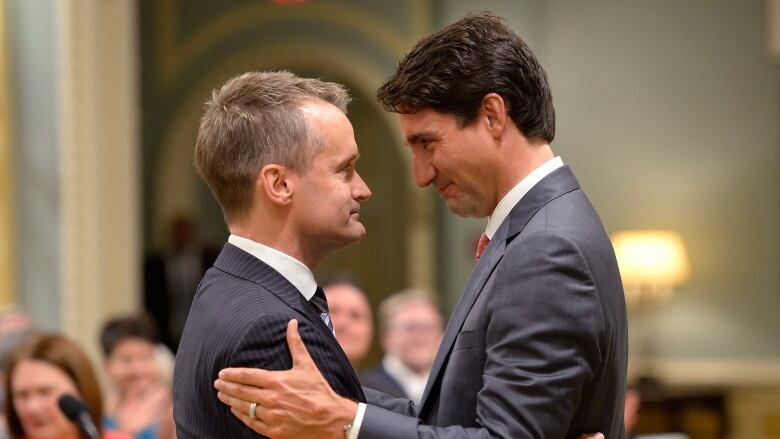 A man with thin blonde hair hugs a man with brown hair wearing a grey suit.