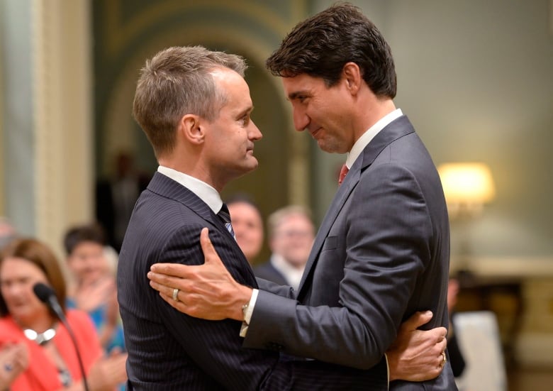 A man with thin blonde hair hugs a man with brown hair wearing a grey suit.