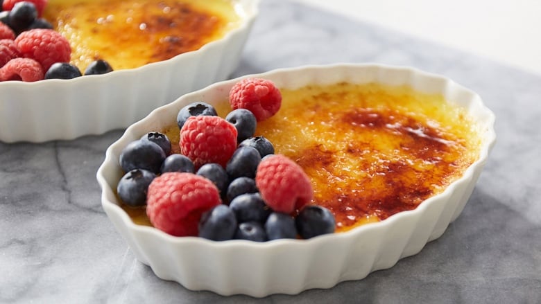 A Crme Brule in a white baking dish on a marble surface. The Crme Brule is topped with raspberries and blueberries. 