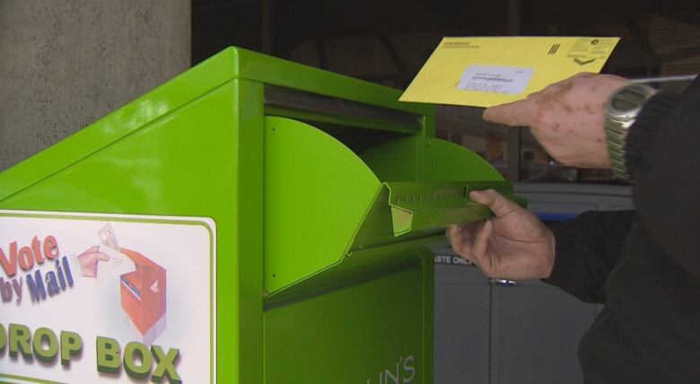 A hand places an envelope in the slot of a green mailbox which says 