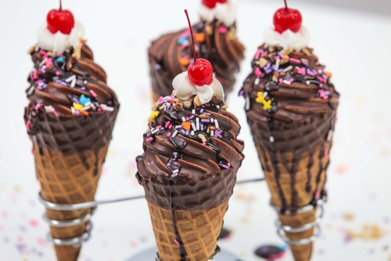 4 ice cream cones in a metal tray. the ice cream is dipped in chocolate and topped with cherries and whipped cream. 