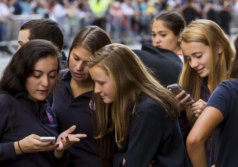 A group of teen girls in school uniforms look at their phones as they stand outdoors.