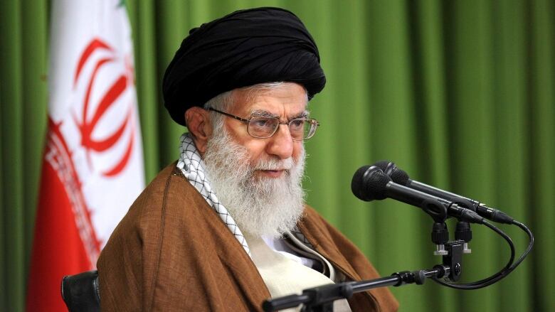 An old man in a turban sits in front of an Iranian flag.