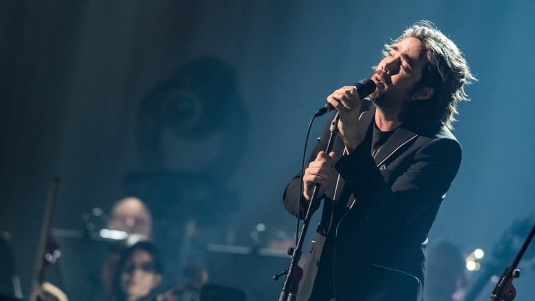 Patrick Watson, a white man with greying hair, sings into a microphone onstage.