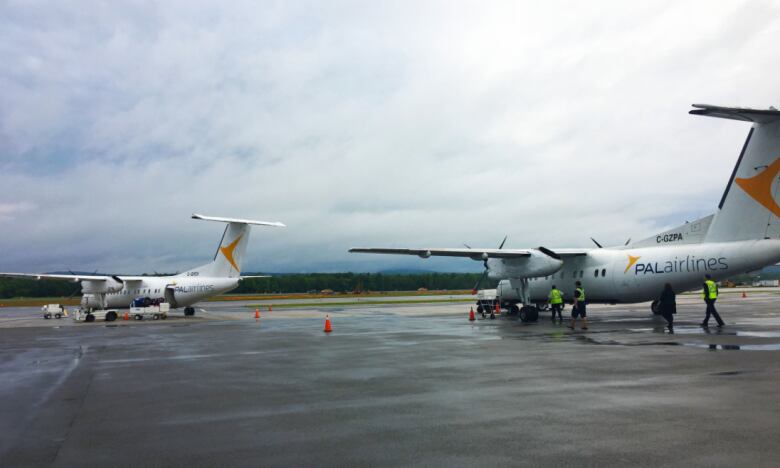 Two passenger planes sit on an airport tarmac, while airport staff with security vests walk around one of them.