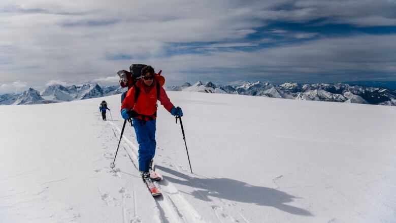 Two cross-country skiers with large packs on a snowy high plateau with mountain peaks in the distance.