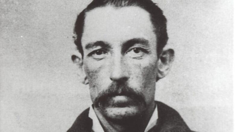 A man with a full mustache and high collar with a loose cravat in an old 1800s-era black and white photo looks straight into the camera with a solemn face.