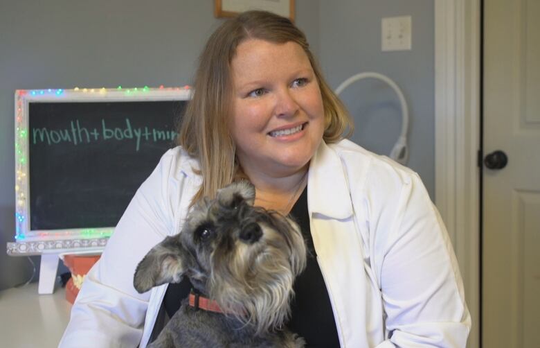 A woman in a white lab coat smiles as her dog sits on her lap.