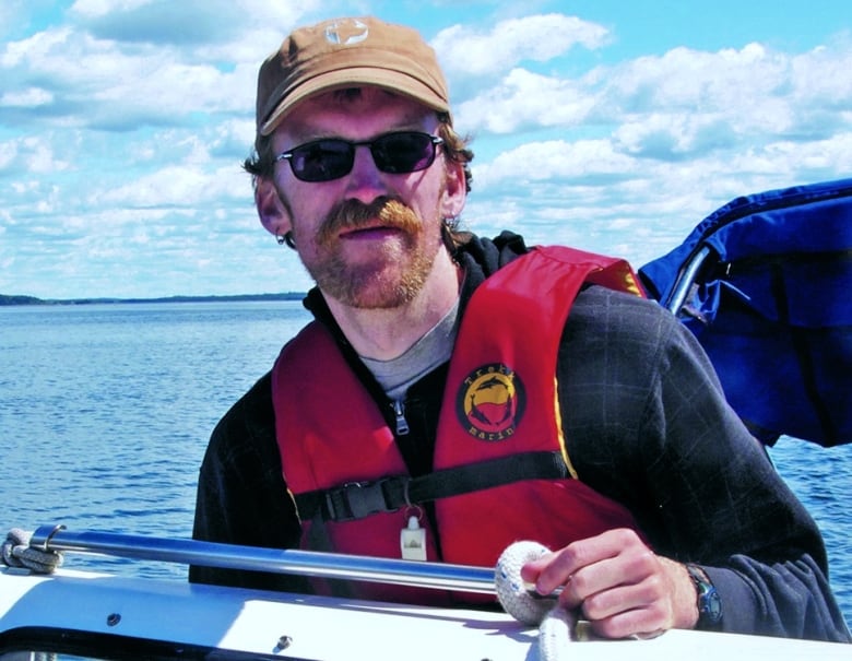 Man, with beard, wearing hat, sunglasses, and life jacket, looking at camera with neutral expression.