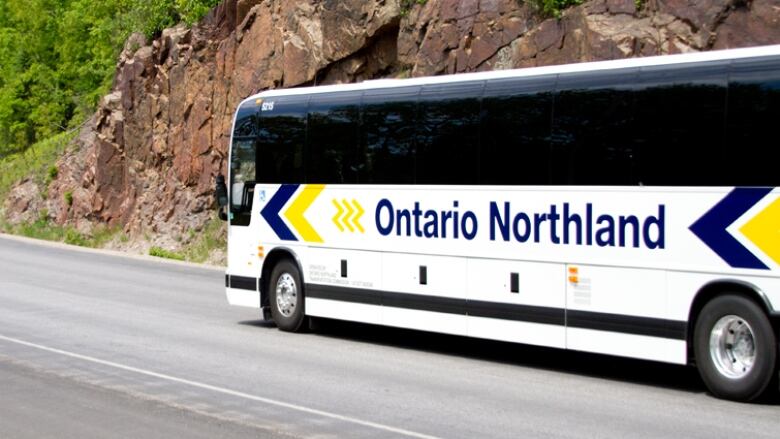 An Ontario Northland bus on the highway passing a rock face and heading in toward a tree-lined stretch of road. 