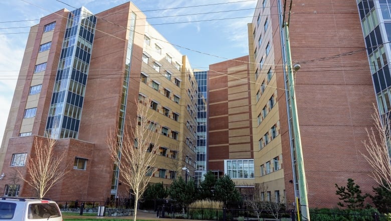 A large red and light brown brick hospital with a line of trees on the property and a few deciduous trees in front of the sidewalk of the building. 