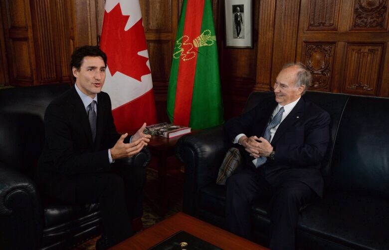 Two men in suits sit talking on a couch.