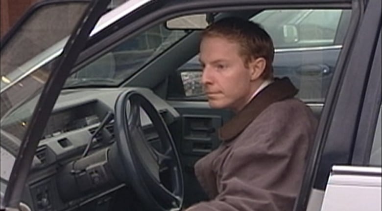 A man with a brown jacket is pictured sitting down on a car behind the steering wheel.