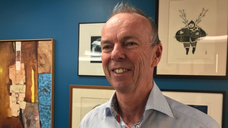 A smiling man wearing a blue button-up shirt stands in front of a brightly painted wall with a gallery of artwork in frames.