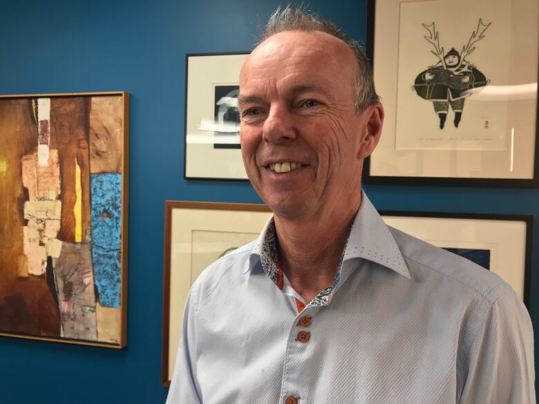 A smiling man wearing a blue button-up shirt stands in front of a brightly painted wall with a gallery of artwork in frames.