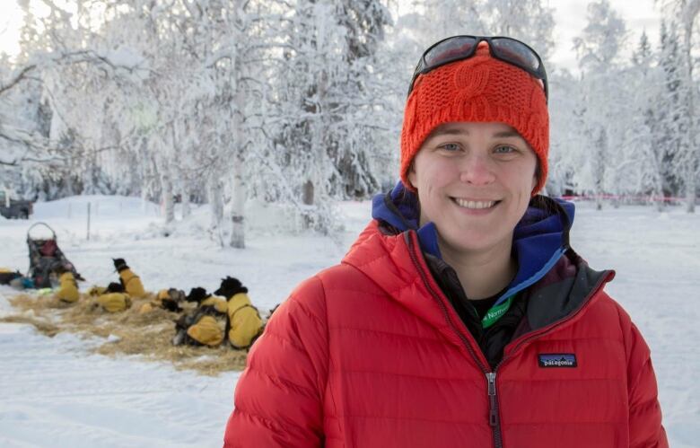 A woman in a red jacket and toque stands in front of sled dogs, lying on hay. 