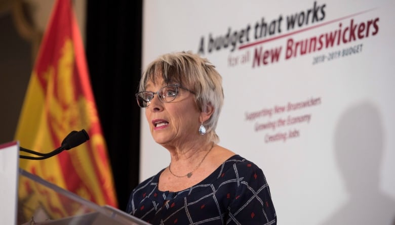 A woman woith grey haor and glasses stands in front of a sign that says A Budget that Works for New Brunswickers.