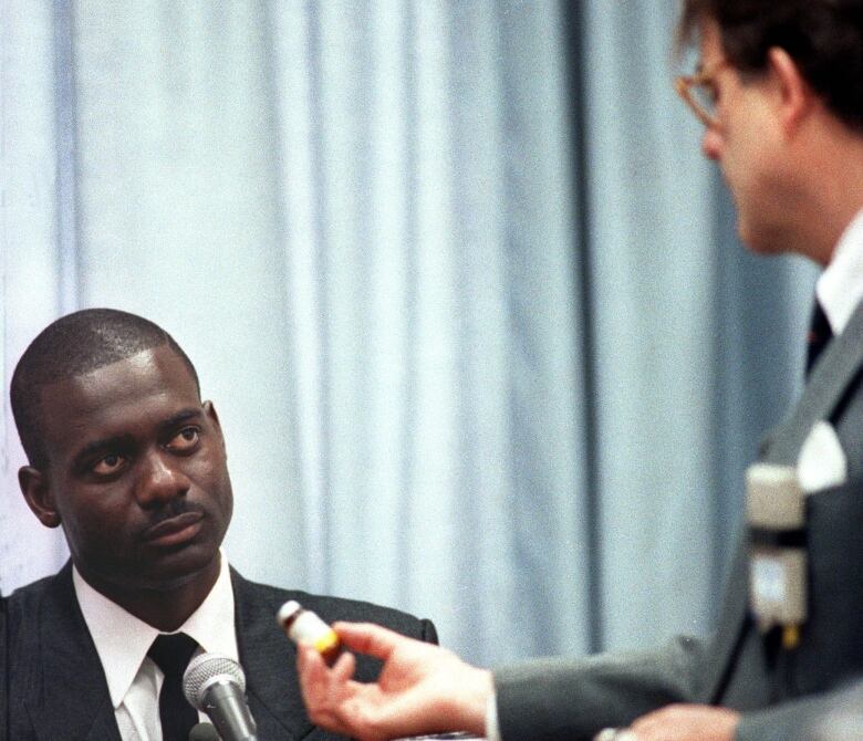 A man in a suit, seated, looks at another man who presents him with a vial.