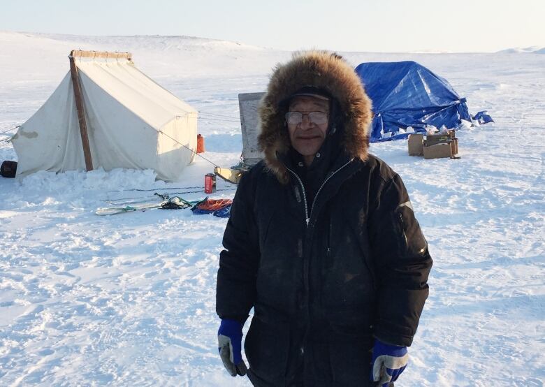 A man in a parka stands outside at a winter campsite.