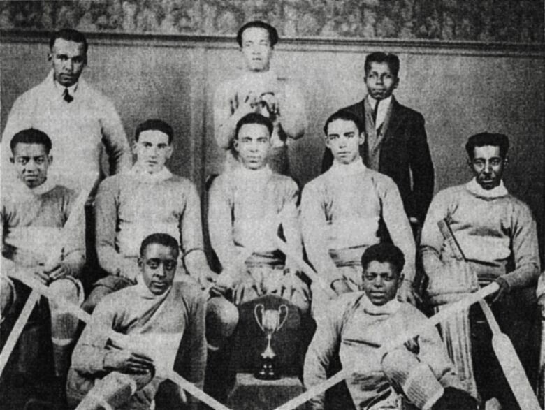 A old picture of a group of Black men wearing old hockey gear sitting around a trophy