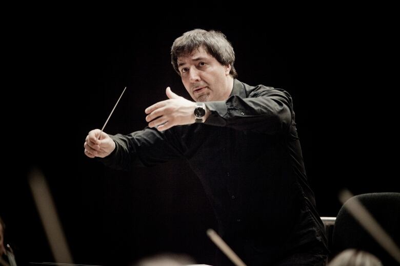 A man with dark hair is shown with his hands in front of him, while conducting an orchestra. The background is dark.