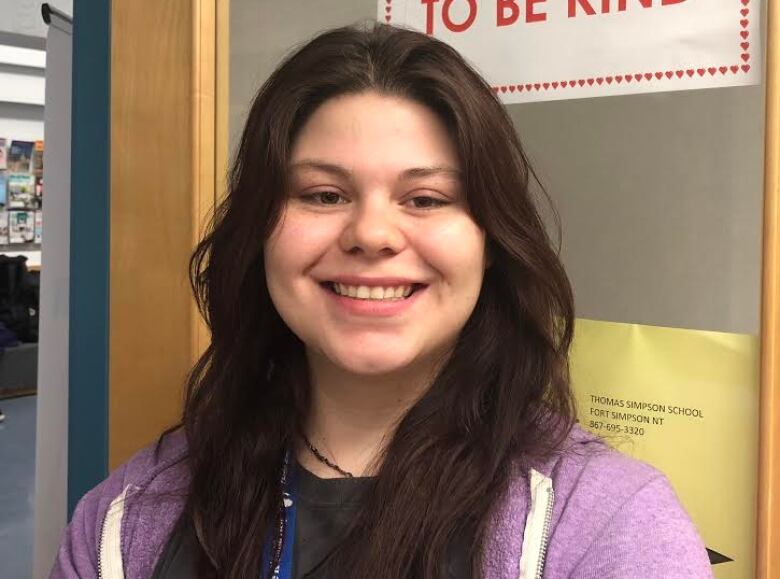 A woman with a purple zip up hoodie and long brown hair looks into the camera. She's standing in front of a door.