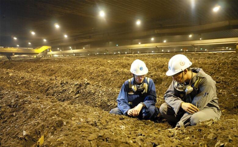 Two men wearing helmets bent down inspecting mounds of dirt-like compost inside a facility. 