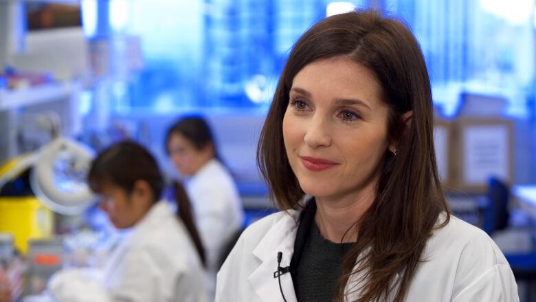 A doctor wearing a white lab coat is in a lab with people working behind her.