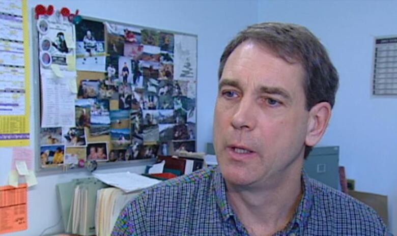 A middle-aged man looks off to the left. Behind him are folders, a filing cabinet and wall-mounted board covered in photos and notes.