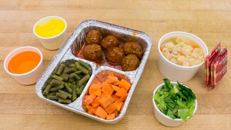 Meal made of meatballs, green beans and carrots in a to go box with two cups of condiments, one bowl of noodle soup and a bowl of salad on the side on a wooden table. 