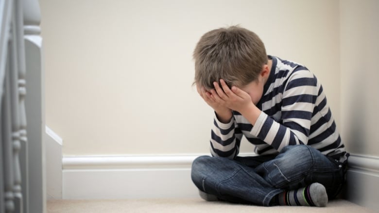 A boy sits in a corner and holds his head in his hands.