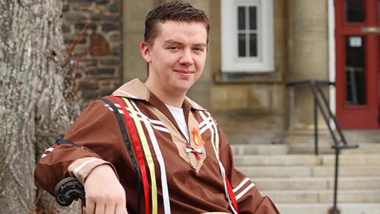 A man sits on a bench and poses for a photo. He has short brown hair. He is wearing a brown shirt with black, red, yellow and white stripes. 