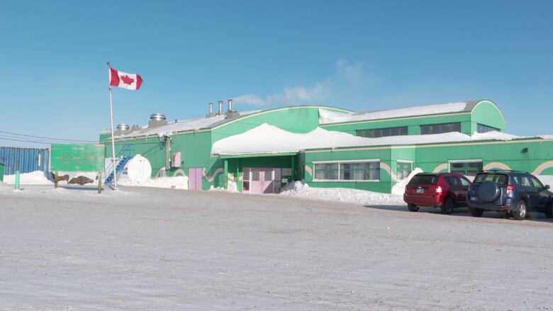 A light green school sits on snowy landscape below a blue sky. In the foreground, a Canadian flag is hoisted.