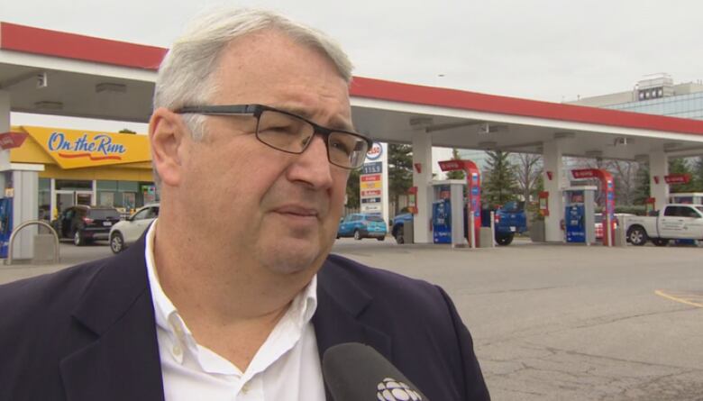 A man with white hair and glasses is seen standing in front of a gas station