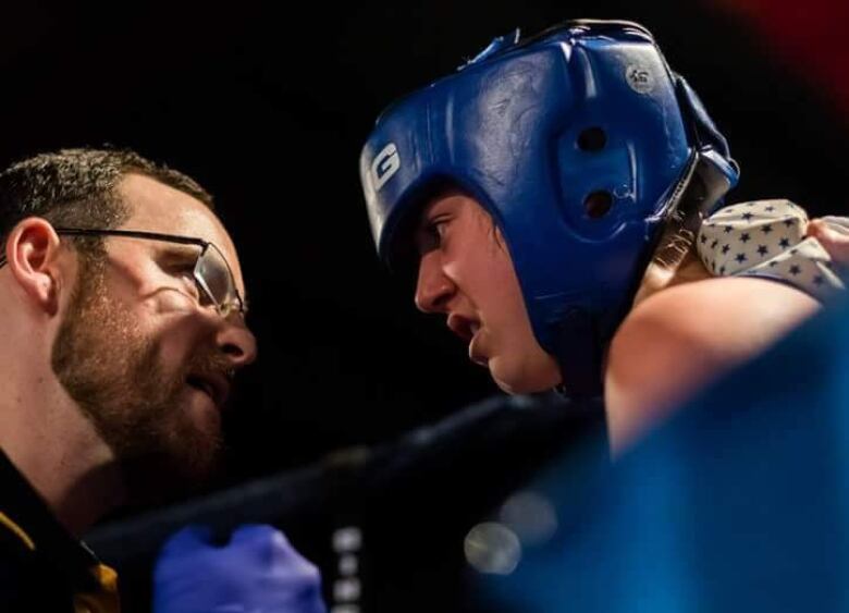 A man with a beard and glasses speaks up close to a female boxer in gear.