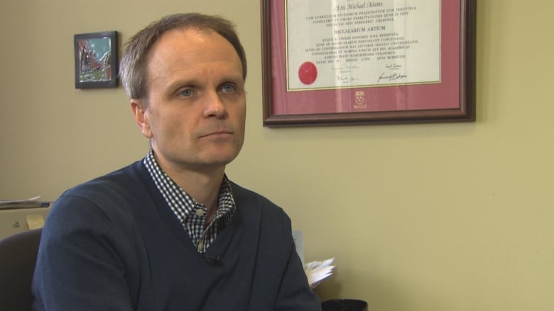 A man looks off to the side of the frame. He's in a beige-painted room with a university document framed on the wall behind him. 