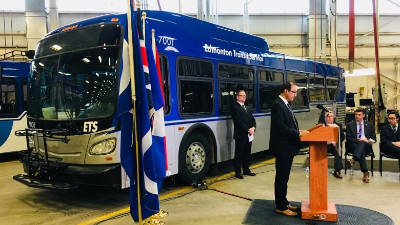  A man in a suit stands at a podium in front of a full-size city transit bus.