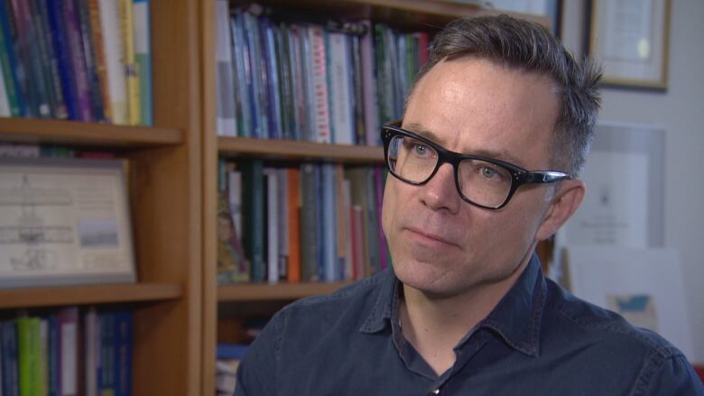 Tim Caulfield looks off camera as he is interviewed. He's sitting in front of shelves lined with books.