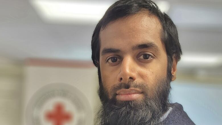 A dark haired man stands indoors looking straight at the camera. The background is slightly out of focus.