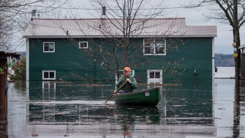 A man in an orange hat paddles a green canoe in floodwaters in front of a green flooded home.