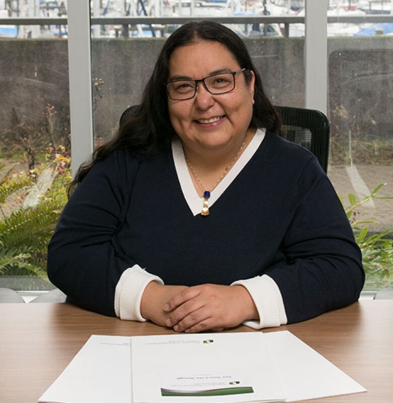 A smiling Indigenous woman with glasses and long dark hair sits at a desk with her hands clasped in front of her.