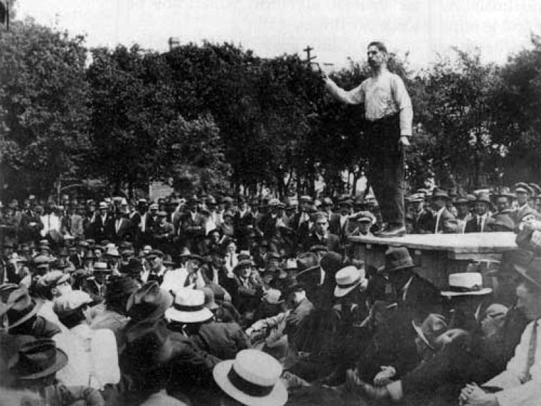 Black and white photo shows a man with short hair and a moustache, He wears suspenders on his pants and stands about a crowd on a platform and he speaks and gestures with his right arm.