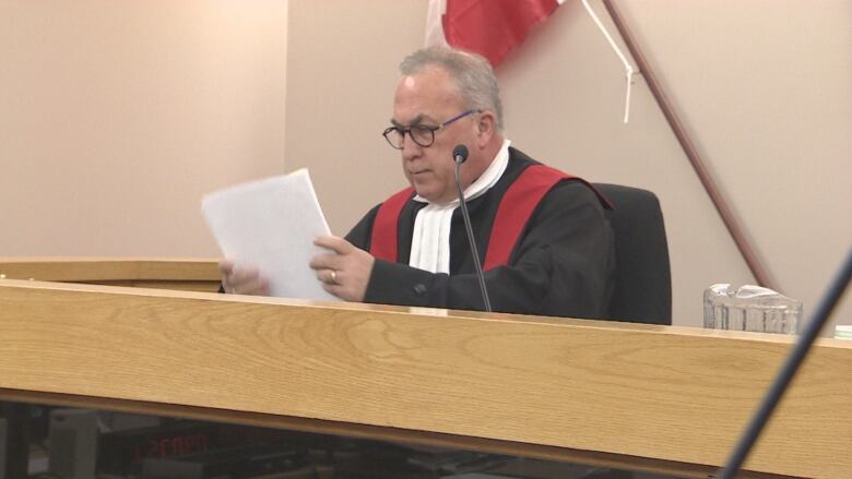 An older white man in a black robe sits behind a bench in a courtroom.