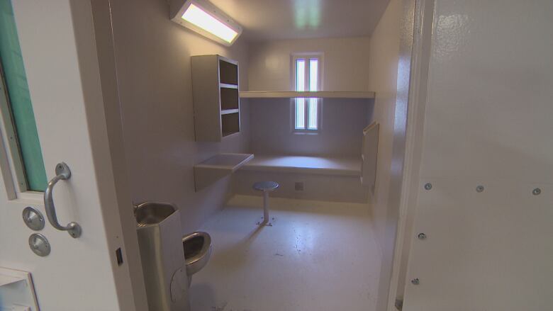 A small room with a metal toilet, table attached to the wall, seat attached to the floor, a bunk bed and a window.