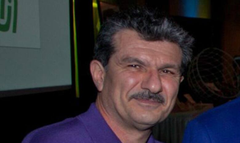 A man with a moustache, wearing a purple shirt, smiles at the camera.