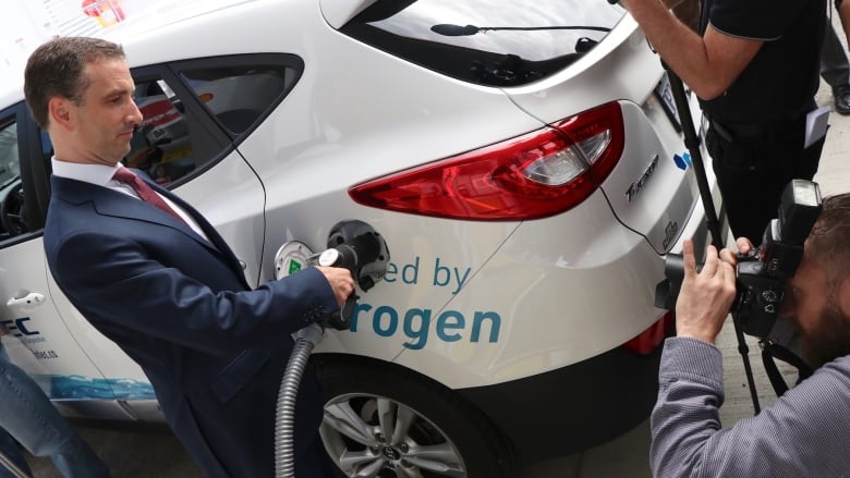 A man in a suit mimes fueling up a hydrogen vehicle at a fueling station in Vancouver while photographers capture the moment.