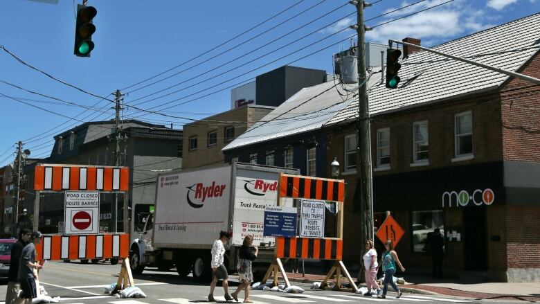 Road signs that say Road Closed are clustered at any intersection in front of businesses. A large Ryder truck is parked behind the signs.